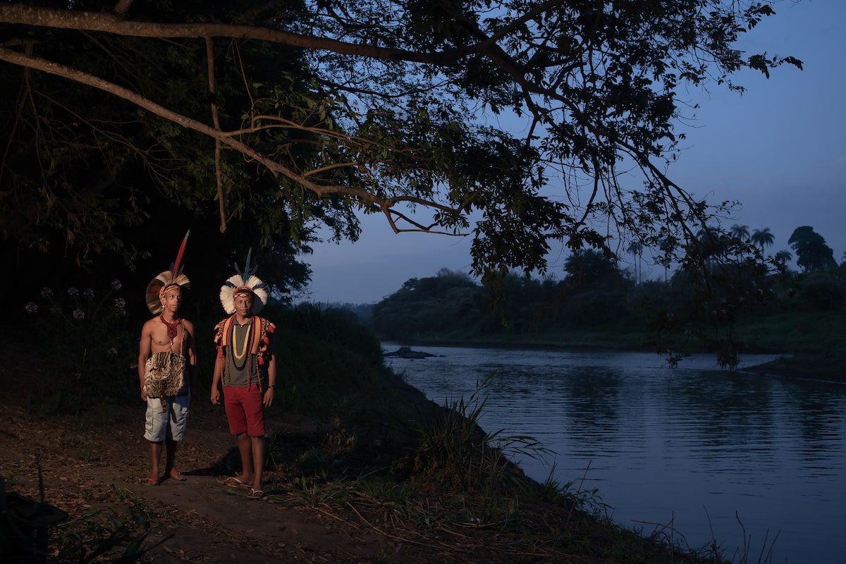 Dois indígenas em pé na margem de um rio ao entardecer.